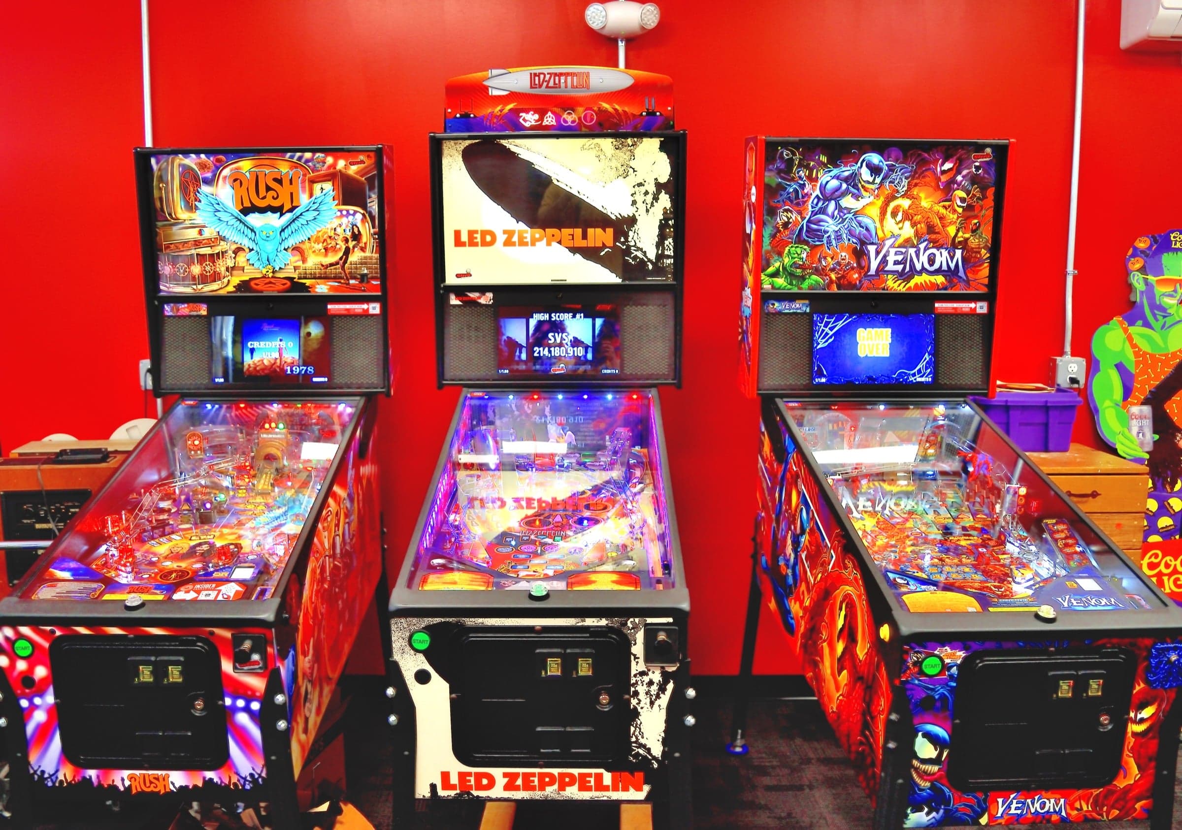 Rush, Led Zeppelin and Venom pinball machines against a red wall in the unfinished room next door