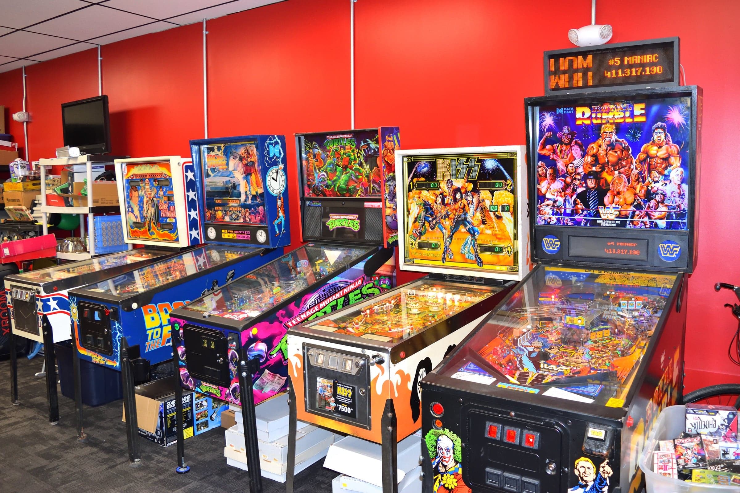 A row of vintage pinball machines against a red wall, including Evel Knievel, Back to the Future, Teenage Mutant Ninja Turtles, KISS and WWF Royal Rumble