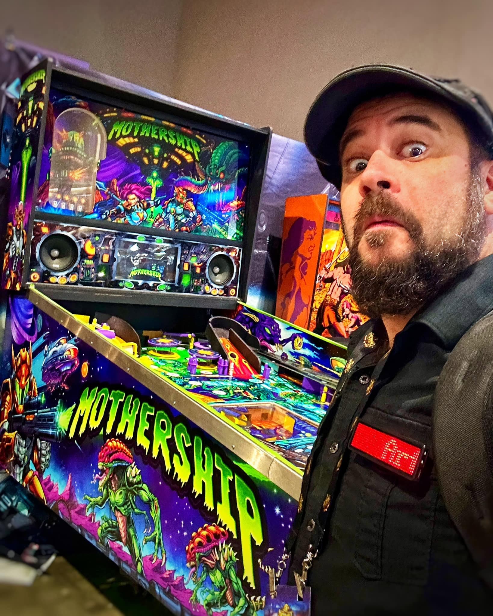 Brian Allen pulling a wide-eyed face beside the Mothership pinball machine, its cabinet and backglass covered in his art of armored soldiers and green alien creatures