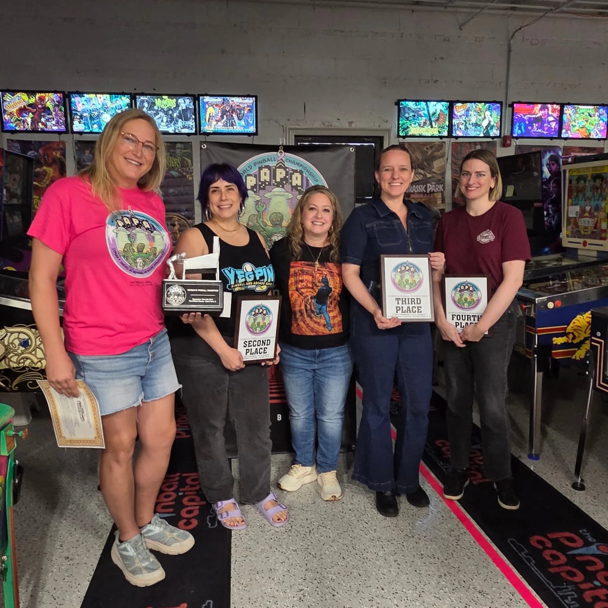The PAPA 2026 Women's finalists holding the trophy and placement plaques in front of a row of machines at The Pinball Capital