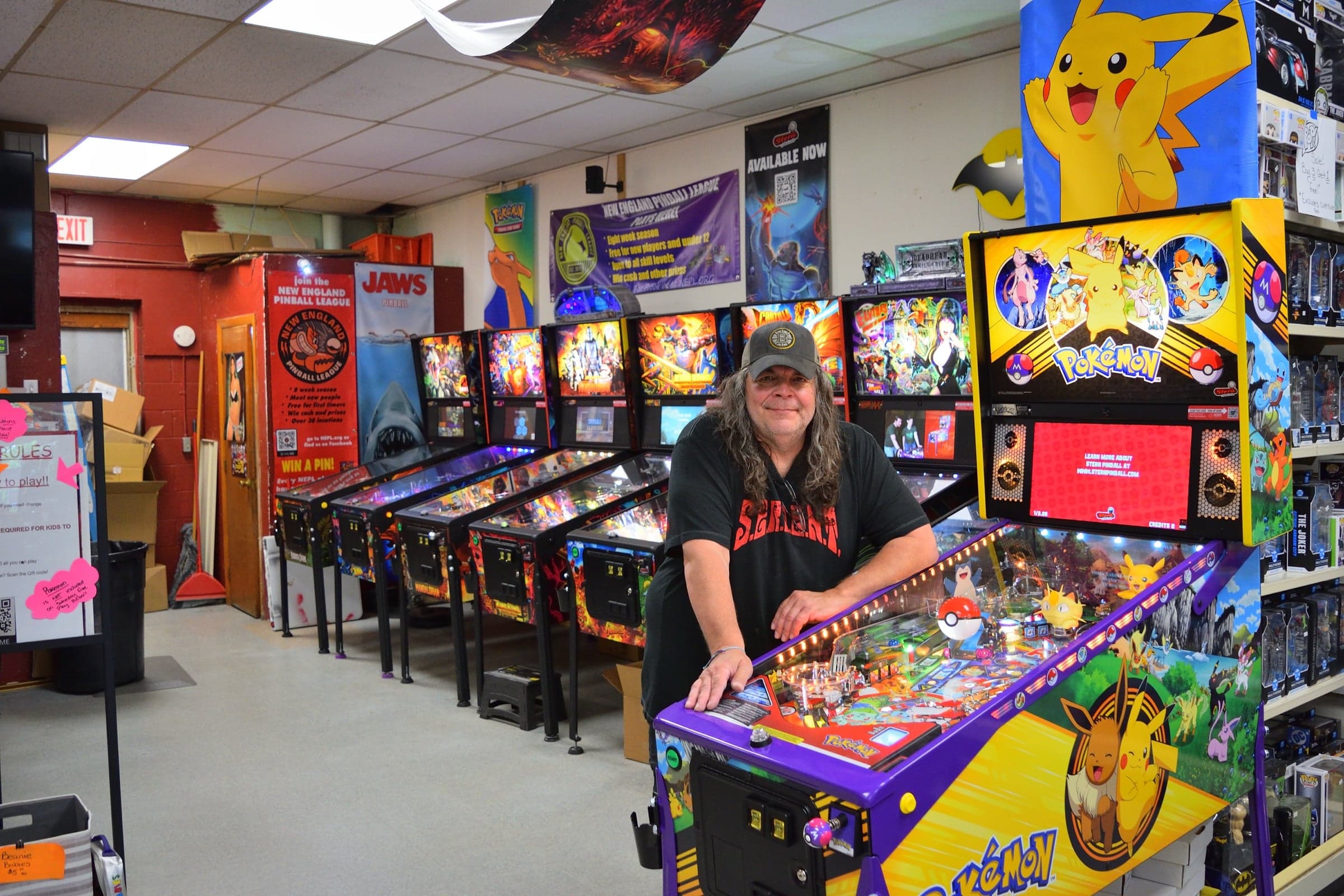 Don Berrios leaning on a Pokemon pinball machine with a row of Stern games and a New England Pinball League banner behind him