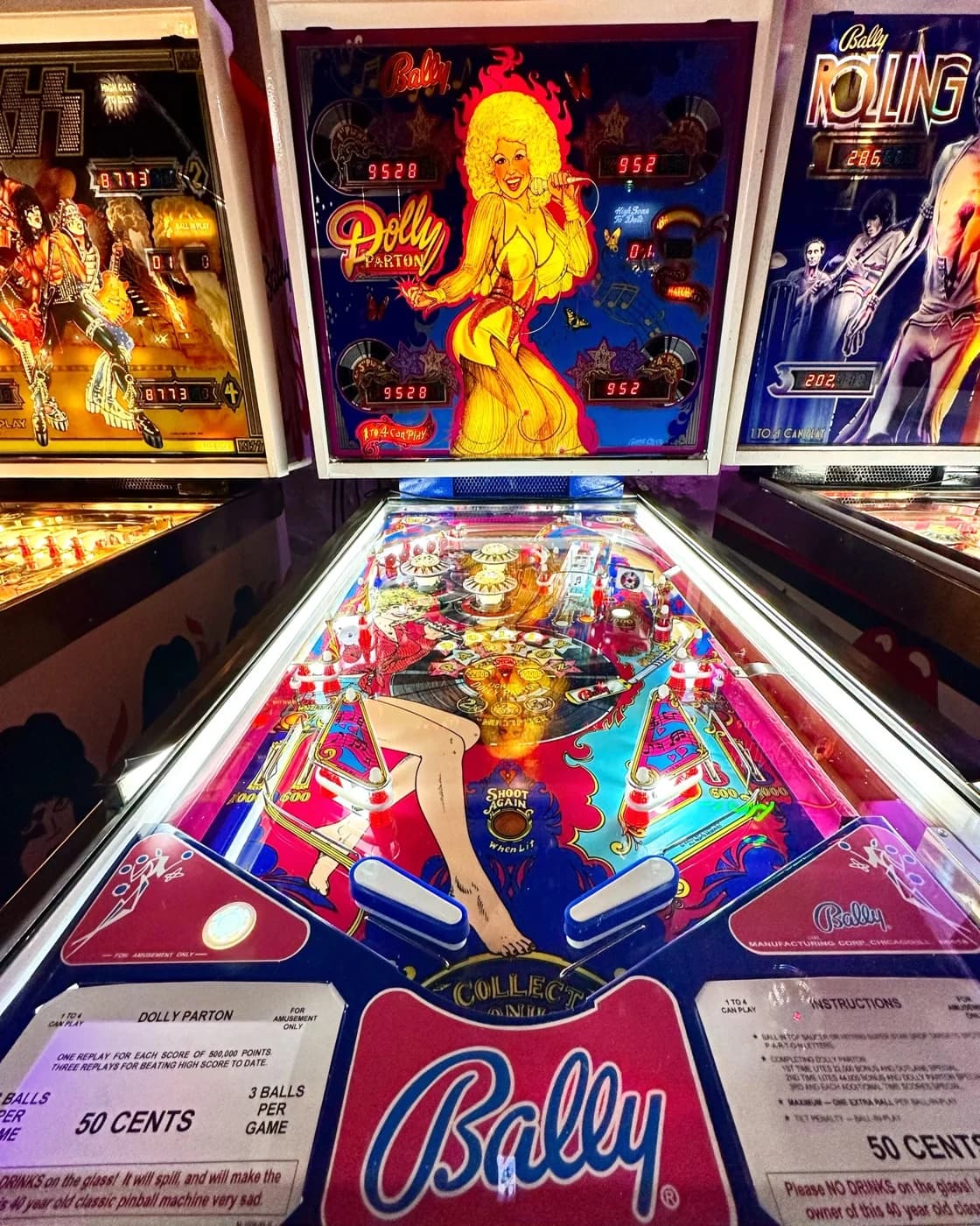 A Bally Dolly Parton pinball machine on location, backglass lit, with the full playfield in view: Parton drawn barefoot in denim across the lower playfield, red and blue pop bumpers and drop targets above, and the apron cards reading 