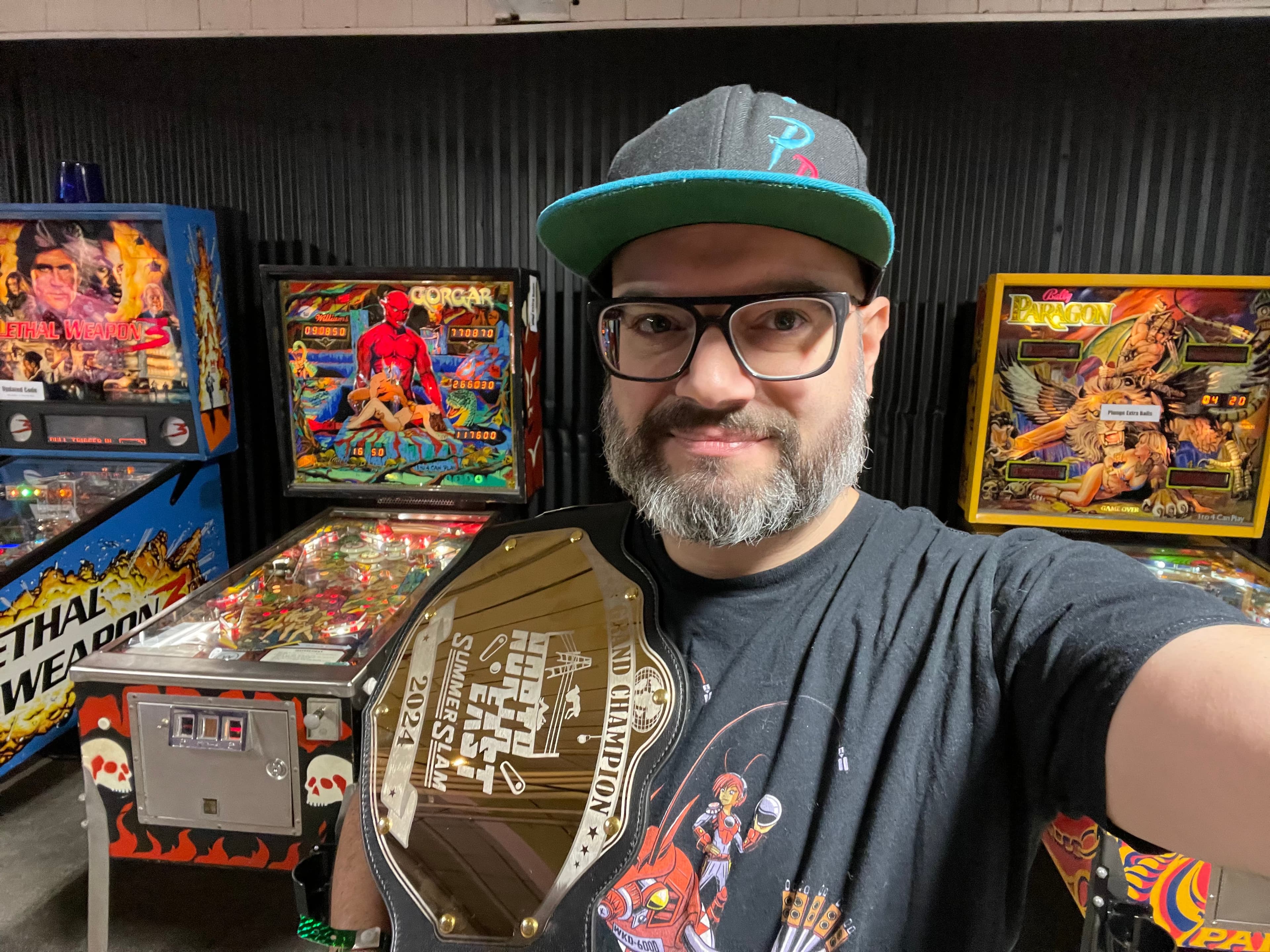 Ed Giardina holding a North East Summer Slam championship belt in front of pinball machines