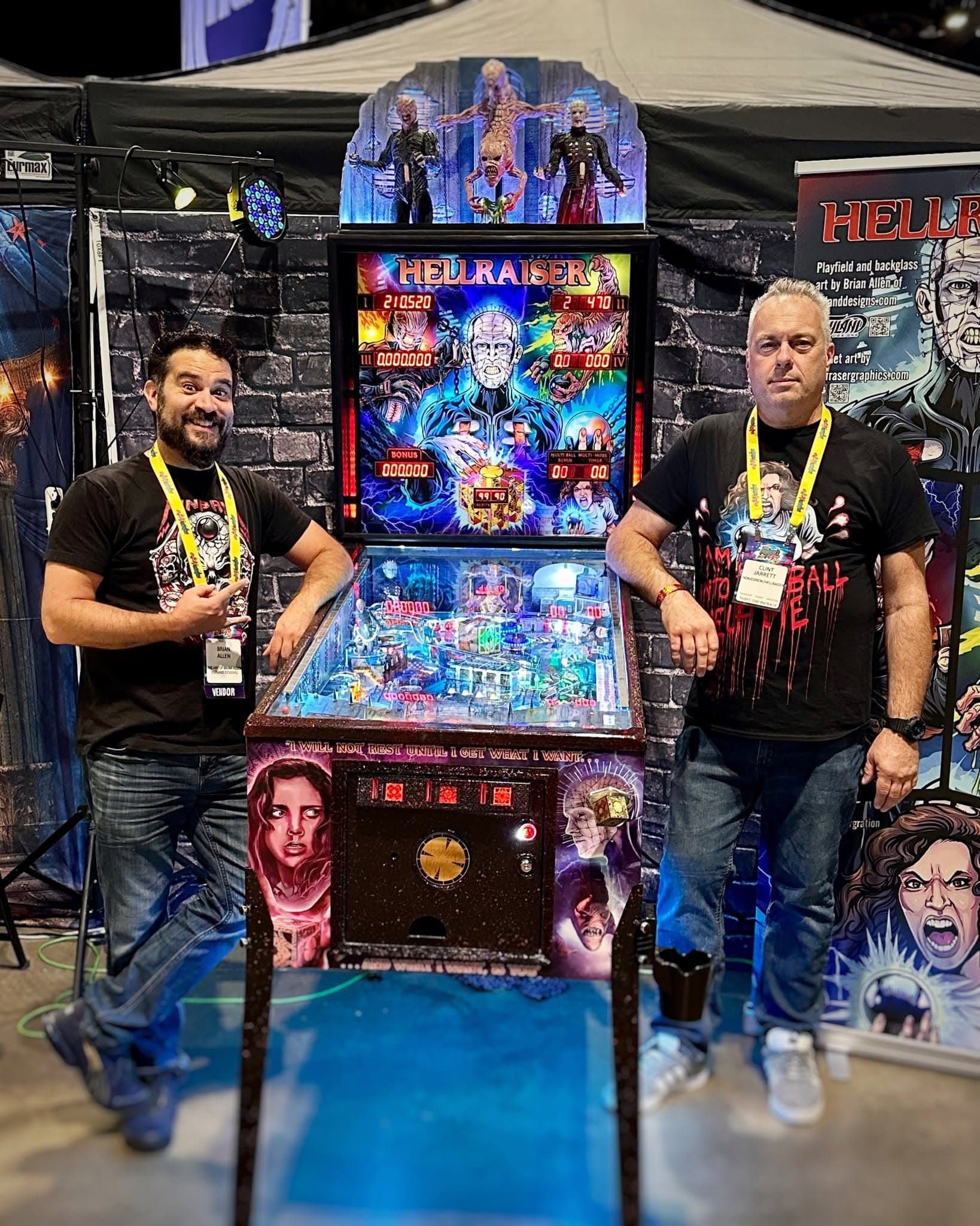 Brian Allen and the game's designer, Clint Jarrett, leaning on either side of the Hellraiser homebrew pinball machine at a convention booth, its backglass showing Pinhead holding the puzzle box
