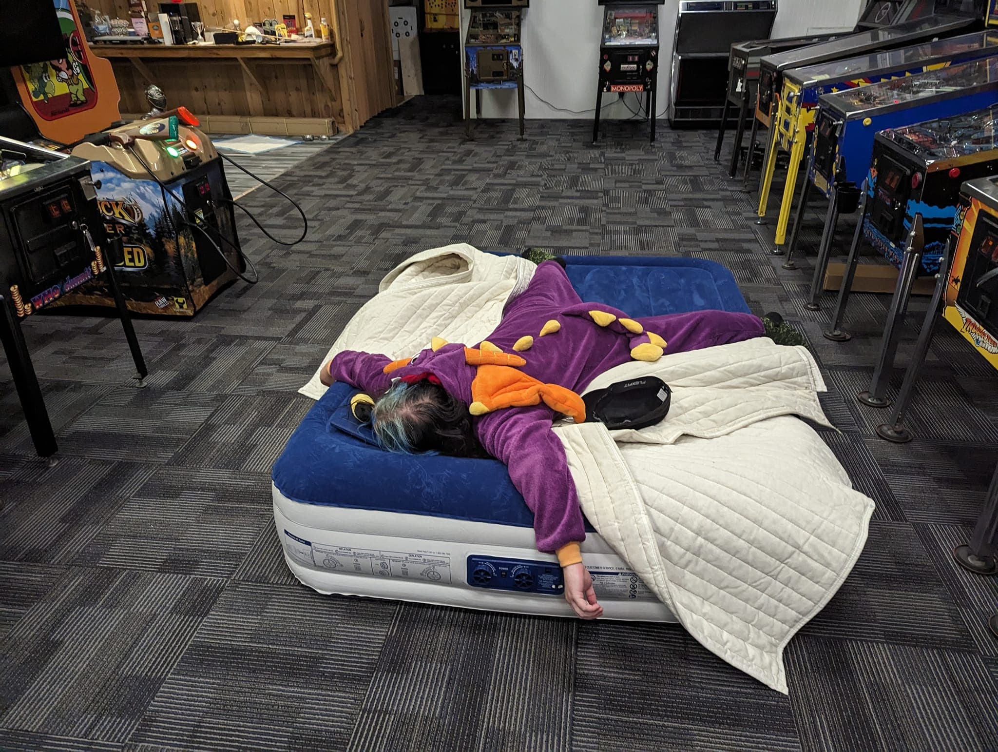 Person in purple costume sleeping on air mattress surrounded by arcade machines