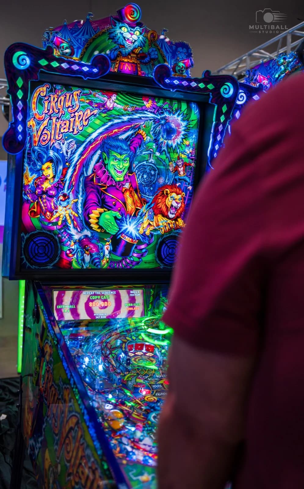 A lit Cirqus Voltaire backbox and playfield at Pinball Expo 2026, a player's shoulder in the foreground