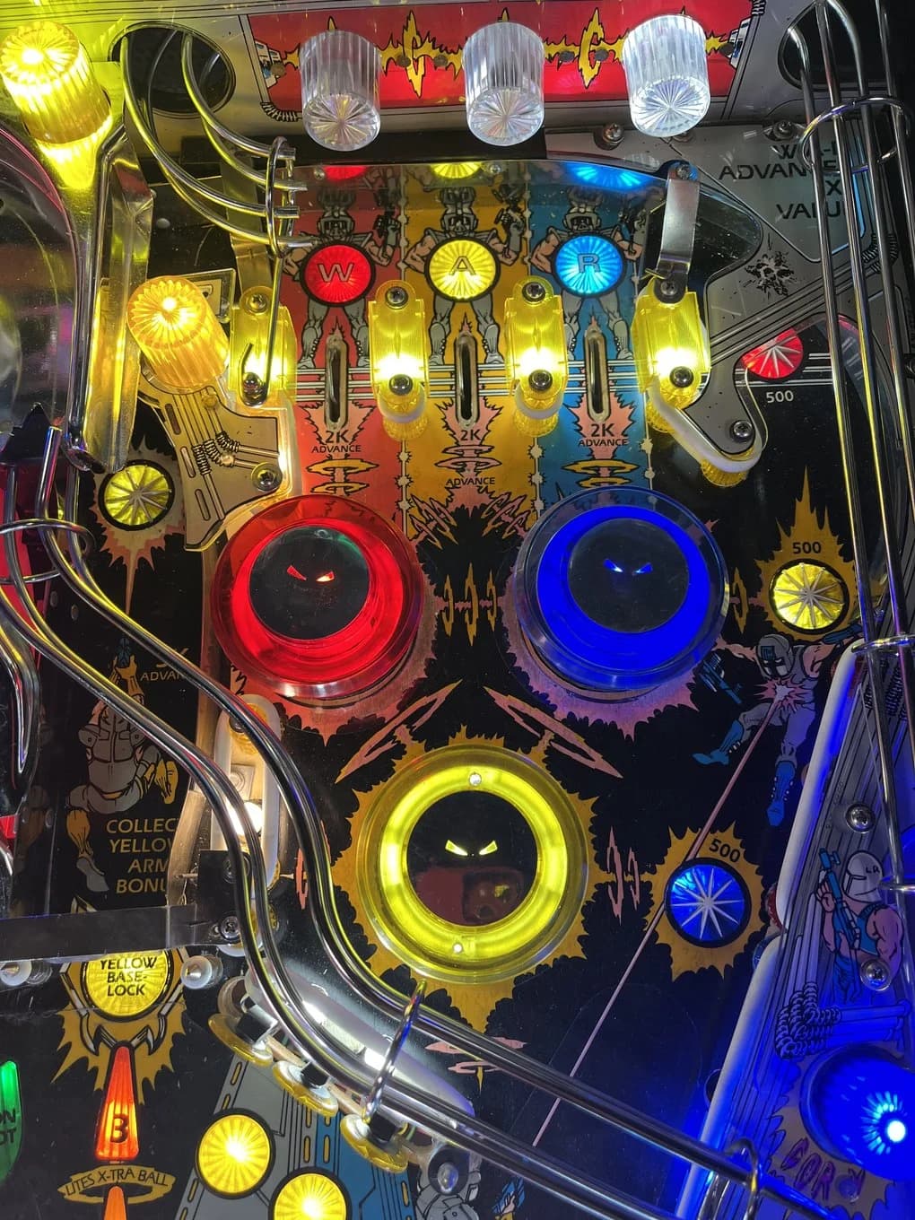 The W-A-R top lanes lit red, yellow and blue with 2K ADVANCE under each, three pop bumpers above them, and the colored saucers below