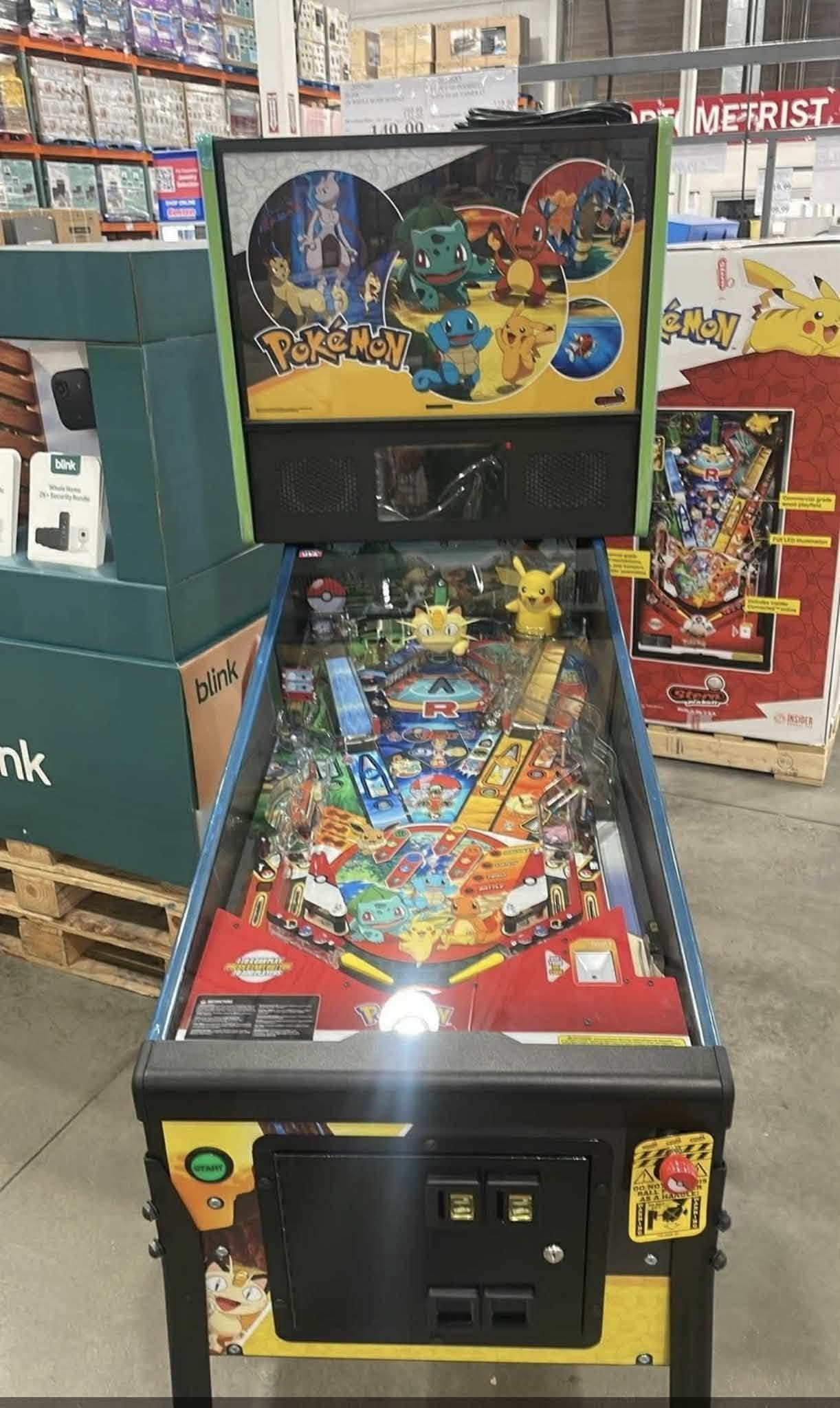 The playfield of the Stern Pokemon Home Edition seen head on, with a Meowth toy at the centre, a Pikachu sculpt at the right and a flat Poke Ball at the upper left, and the machine's retail box standing alongside