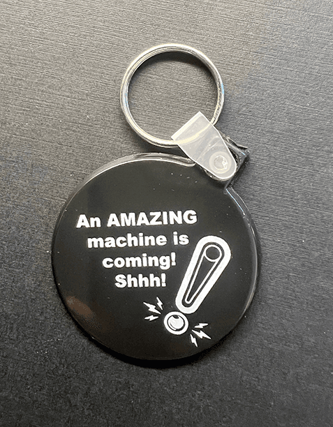 Black keychain with humorous text: An AMAZING machine is coming! Shhh!