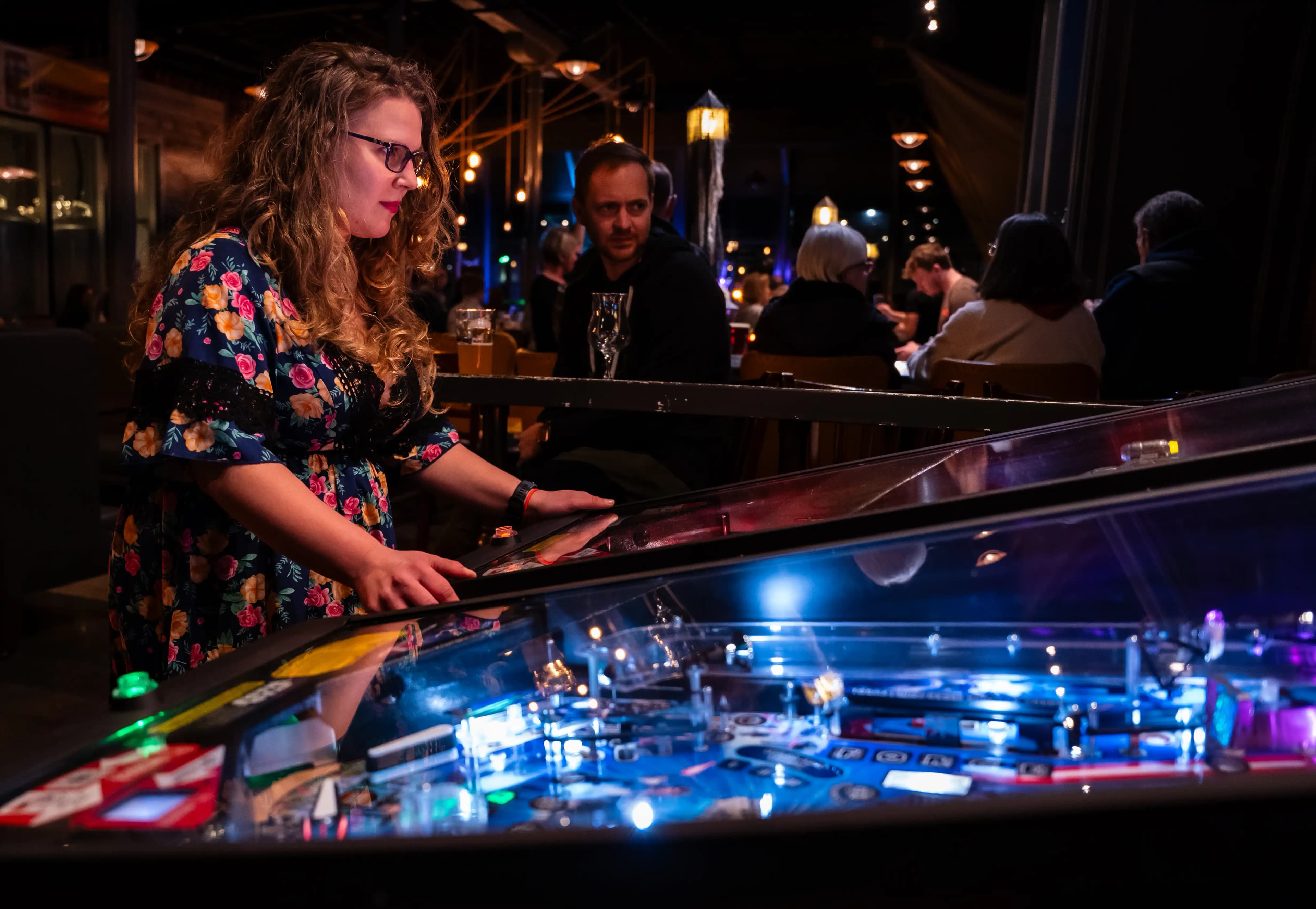 Woman playing pinball machine in dimly lit bar with colorful lights