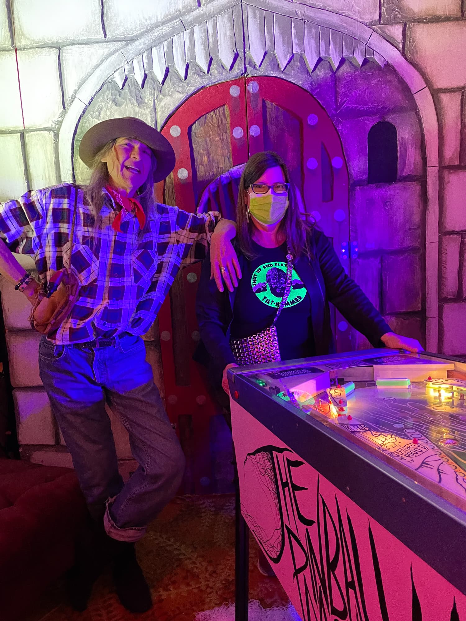Two people posing next to a colorful pinball machine with purple lighting